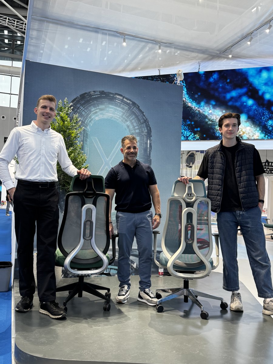 Gustas Smulkstys and Lukas Grigalis with the ALEGRE Design designer beside two chairs at a fair stand